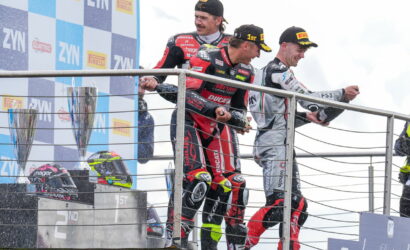 Three motorcycle racers in racing suits celebrate on the podium with trophies on the left and helmets on the railing.