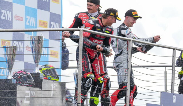 Three motorcycle racers in racing suits celebrate on the podium with trophies on the left and helmets on the railing.