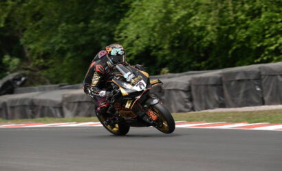 Rider in black and orange racing gear leans into a tight turn on a race track with safety barriers in the background, motorcycle at speed.