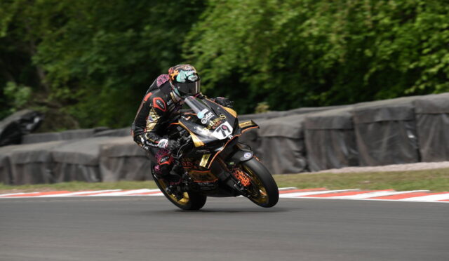 Rider in black and orange racing gear leans into a tight turn on a race track with safety barriers in the background, motorcycle at speed.