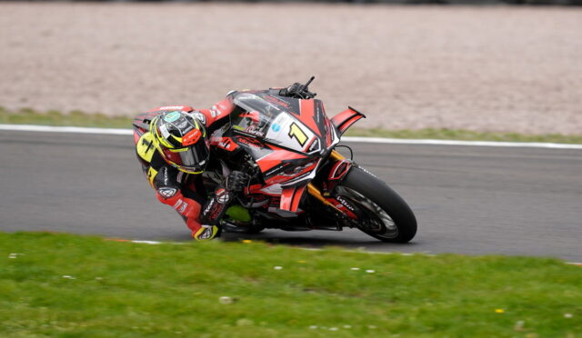 Motorcycle racer in red and black leans into a track turn on a racing bike with number 1 on the front helmet visor shows yellow accents