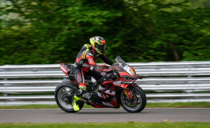 Motorcycle racer in red and black suit riding a red racing bike on a track with green trees in the background, number 1 visible on the bike's front.