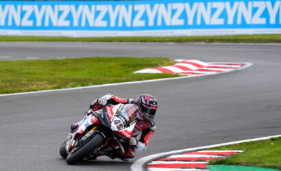 Motorcycle racer in red and white gear leaning into a sharp turn on a race track; bike number 41 with sponsor logos, blue ad banners in the background.