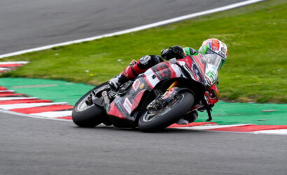 Moto racer in red-black gear leans into a tight corner on a race track, bike tilted with red/white curbs nearby.