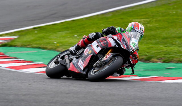 Moto racer in red-black gear leans into a tight corner on a race track, bike tilted with red/white curbs nearby.