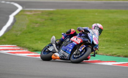 MotoGP rider in blue and pink suit leaning into a sharp turn on a race track; motorcycle heavily tilted with sponsor stickers visible on the fairing.