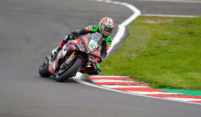Motorcycle racer leaned into a sharp curve on a race track, wearing green, red, and black gear with a colorful helmet.