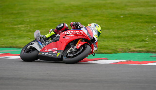 Red Honda sportbike rider leaning into a left-hand turn on a race track, helmeted rider in neon-yellow gear. on-track curbs are red, white, and green, with grass in the background.