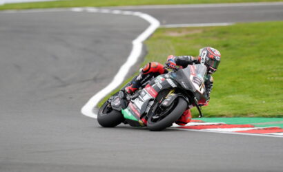 Motorcycle racer leans into a sharp turn on a race track, full racing gear and sponsor decals visible on the bike and suit.
