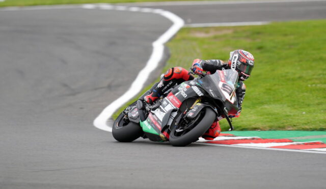 Motorcycle racer leans into a sharp turn on a race track, full racing gear and sponsor decals visible on the bike and suit.
