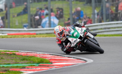 Motorcycle racer leaning into a sharp turn on a race track, red-and-white curb in foreground and blurred spectators in the background.