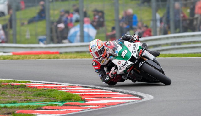 Motorcycle racer leaning into a sharp turn on a race track, red-and-white curb in foreground and blurred spectators in the background.