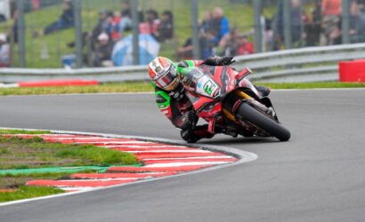 Rider on a red race bike leaning into a curve on a racetrack with red-and-white curbs and blurred spectators in the background.