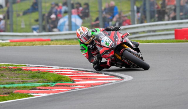 Rider on a red race bike leaning into a curve on a racetrack with red-and-white curbs and blurred spectators in the background.