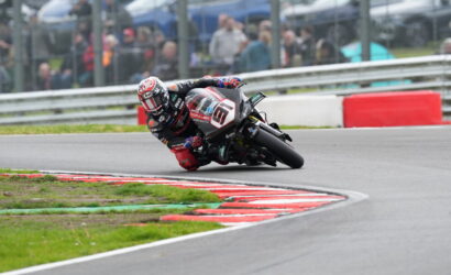 Motorcycle racer leaning into a sharp turn on a race track, spectators blurred in the background behind barriers.