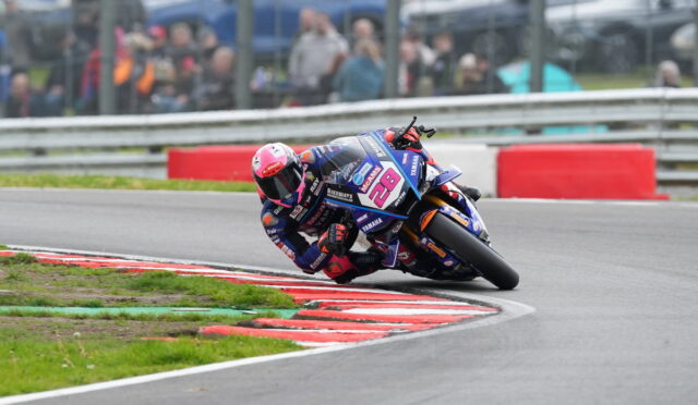 Motorcycle racer leaning into a turn on a racetrack, wearing pink helmet and blue Yamaha livery with number 28, spectators in the background