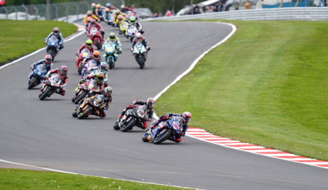 Group of motorcycle racers leaning into a curve on a race track, colorful leathers and bikes, spectators in the background.
