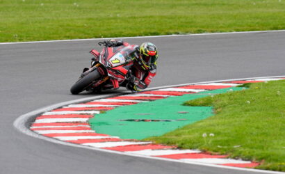 Moto racer in red/black gear leans into a sharp corner on a race track, bike number 7 on red-and-black livery.