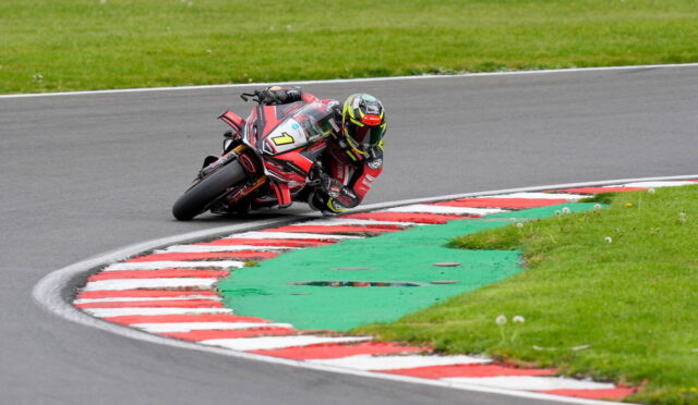 Motorcycle racer in red and black racing suit leaning into a left-hand corner, bike number 7 on a track with red-and-white curbs and green runoff grass behind.