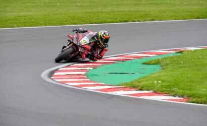 Motorcycle racer in red and black gear leaning into a corner on a racetrack, close to red-and-white curbs.
