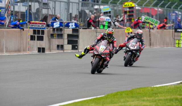 Two sport motorcycles lean into a curve on a race track, rider #7 in front and rider #91 close behind, with barriers and spectators in the background.