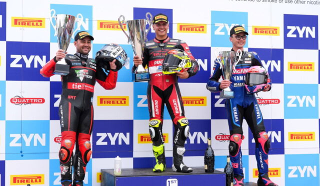 Three motorcycle racers stand on a podium holding large trophies, with the first-place rider in the center wearing red/black Ducati gear, flanked by riders in red/black and blue Yamaha gear in front of a sponsor-filled backdrop.