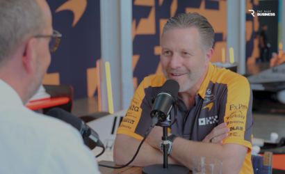 Interview setup: man in orange racing polo speaks into a microphone across from another listener in a studio with branded backdrop and a 'The Race Business' logo in the corner.