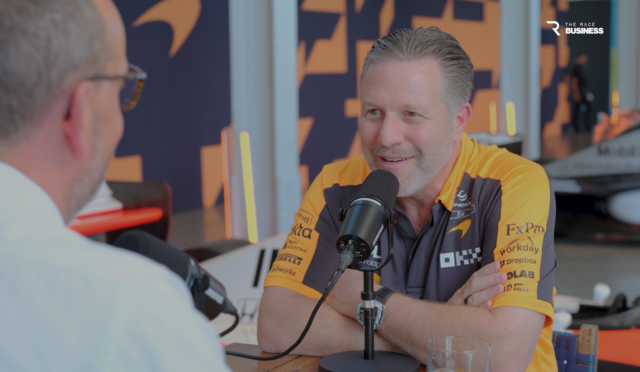 Interview setup: man in orange racing polo speaks into a microphone across from another listener in a studio with branded backdrop and a 'The Race Business' logo in the corner.