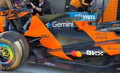 Orange and black Formula 1 car in the pit area with a cameraman and reporter nearby capturing footage with a Sky Sports microphone nearby spectators/crew in the background.