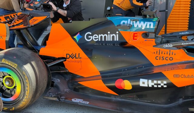 Orange and black Formula 1 car in the pit area with a cameraman and reporter nearby capturing footage with a Sky Sports microphone nearby spectators/crew in the background.