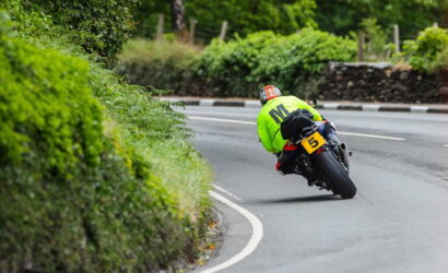 Motorcyclist in a neon green jacket leans into a curve on a winding road with green hedges on the left and a yellow number plate 5 on the bike