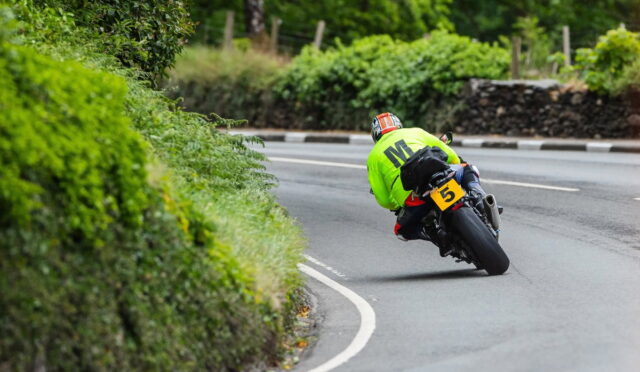 Motorcyclist in a neon green jacket leans into a curve on a winding road with green hedges on the left and a yellow number plate 5 on the bike