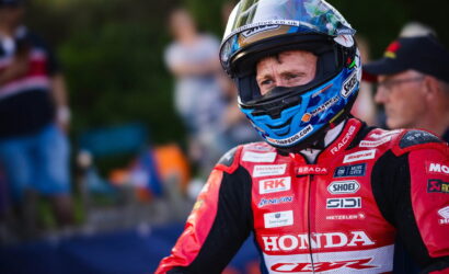 Motorsport rider in red Honda racing suit and blue helmet, facing forward on a track with blurred spectators in the background.