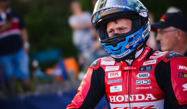 Motorsport rider in red Honda racing suit and blue helmet, facing forward on a track with blurred spectators in the background.