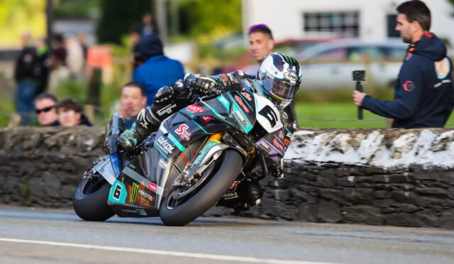 Motorcycle racer in full protective gear leans into a sharp turn on a sponsor-laden racing bike, with spectators and a stone barrier in the background.