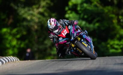 Motorcycle racer in black-puchsia-blue livery leaning into a sharp left turn on a racetrack, helmeted and focused as tires grip the asphalt.
