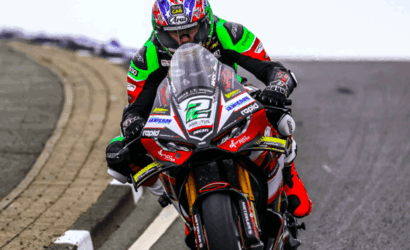 Motorcycle racer in neon green, red, and black leathers leans into a turn on a race track; bike number 21 is visible with sponsor decals.