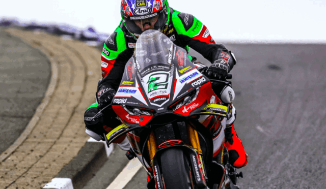 Motorcycle racer in neon green, red, and black leathers leans into a turn on a race track; bike number 21 is visible with sponsor decals.