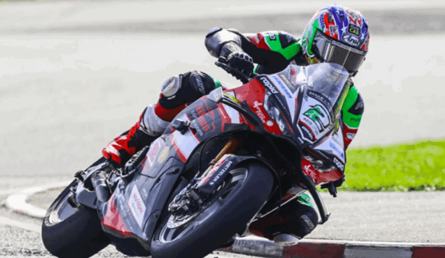 Motorcycle racer in colorful gear leaning into a sharp turn on a track, body tucked close to the bike for speed.