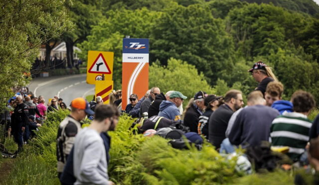 Crowd of spectators along a winding rural road, sitting and standing by hedges with road signs and an orange Hillberry banner in the background.