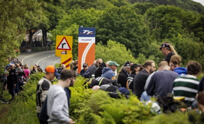 Crowd of spectators sitting and standing along a grassy verge beside a winding road, with racing banners in the distance.