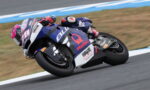 Motorcycle racer leaning into a hairpin on a blue racing bike with number 28 on the front, on a track surface with blue and white curbs.