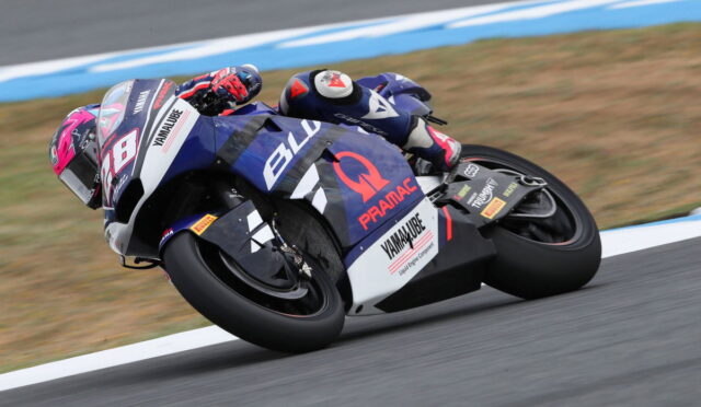 Motorcycle racer leaning into a hairpin on a blue racing bike with number 28 on the front, on a track surface with blue and white curbs.
