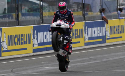 MotoGP rider in a blue-red suit lifts front wheel on a race bike during a wheelie on the track beside Michelin barriers.