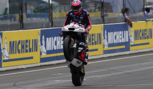 MotoGP rider in a blue-red suit lifts front wheel on a race bike during a wheelie on the track beside Michelin barriers.