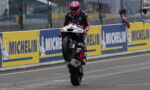 Rider performing a wheelie on a racing bike in the pit lane, with yellow Michelin boards along the barrier behind.