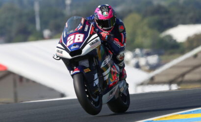 Moto racer in a blue Yamaha bike, pink-helmeted rider leaning into a high-speed turn on a race track.