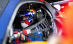 Race car driver in a blue suit and Red Bull helmet sits in cockpit, strapped into a safety harness.