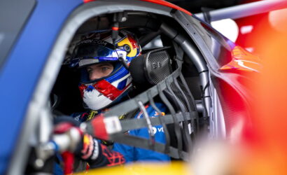 Race car driver in a blue suit and Red Bull helmet sits in cockpit, strapped into a safety harness.