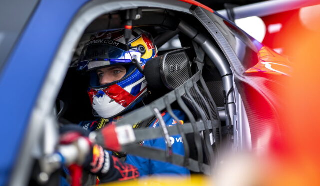 Race car driver in a blue suit and Red Bull helmet sits in cockpit, strapped into a safety harness.